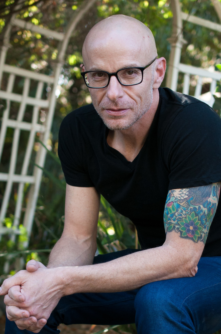 Author David Treuer sits before a garden arbor in a black t-shirt, black glasses, and jeans, with his elbows on his knees and hands clapsed 
