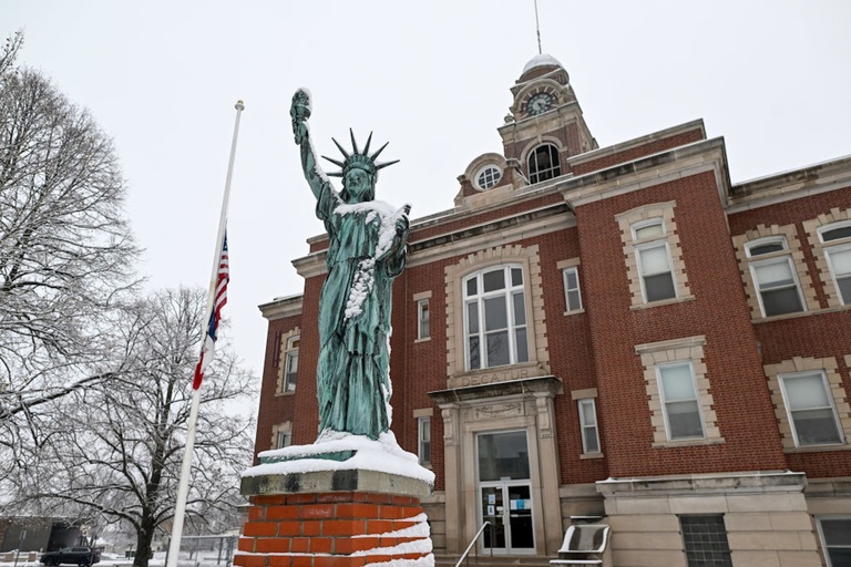 A green-gray statue is lightly covered in snow, standing in front of a brick building with a tower.