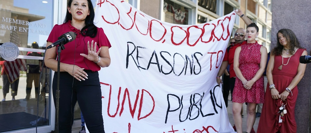 A woman in red speaking in front of a homemade sign about school funding