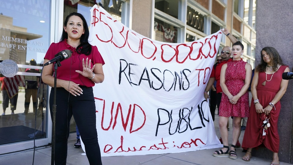 A woman in red speaking in front of a homemade sign about school funding